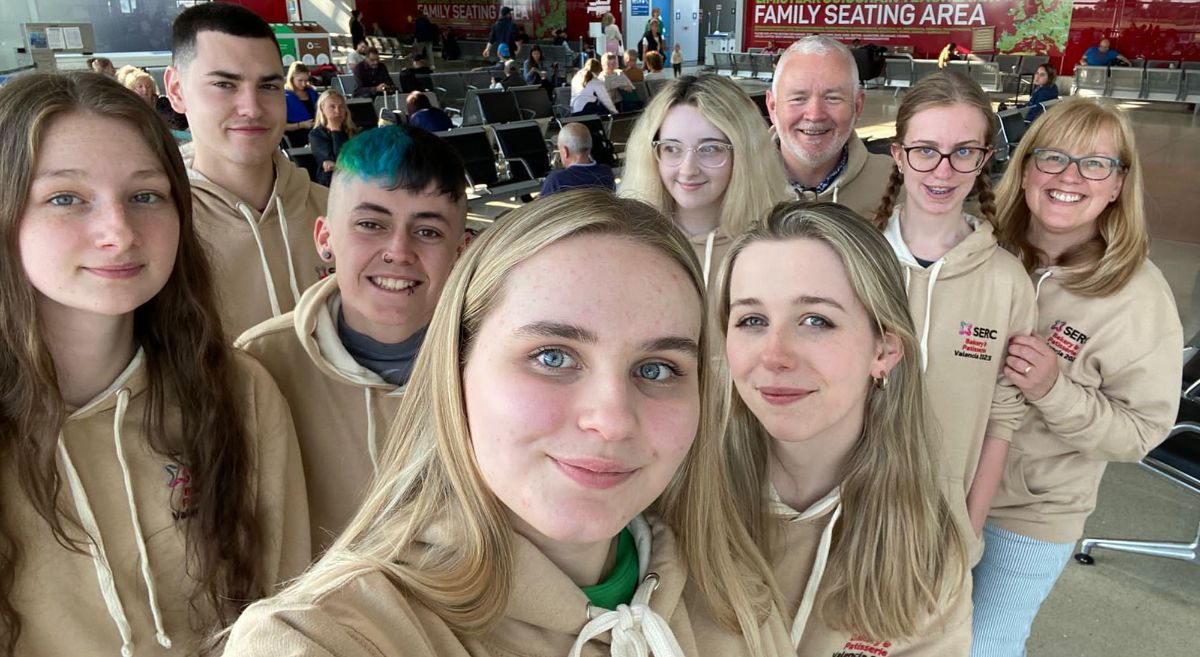 Selfie style photo of a group of  seven students and 2 lecturers all wearing beige hoodies with airport lounge in background